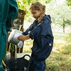 A young girl in an Originals Waterproof Puddle Suit Navy is pouring water from a metal pot into a black bucket, outdoors.